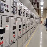 Sala de controle elétrico com uma longa fileira de painéis e equipamentos de monitoramento.
