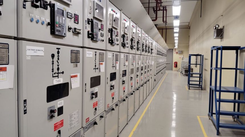 Sala de controle elétrico com uma longa fileira de painéis e equipamentos de monitoramento.