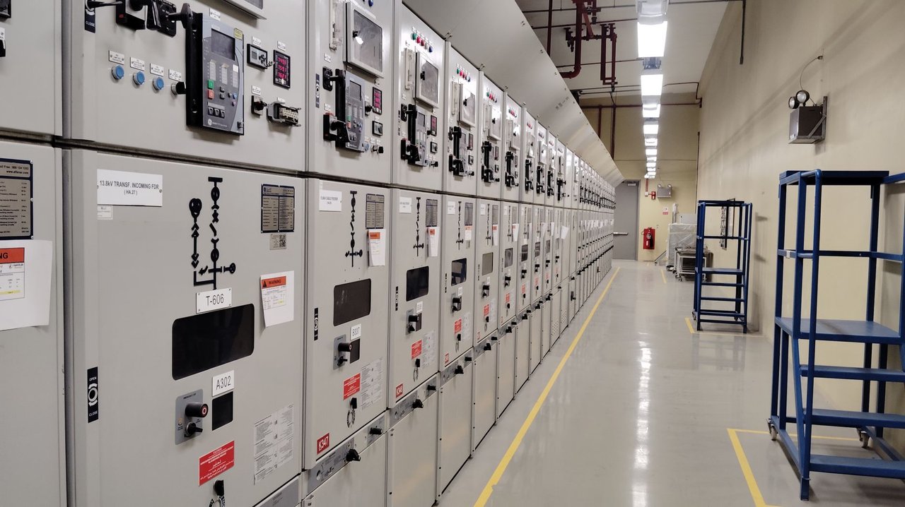 Sala de controle elétrico com uma longa fileira de painéis e equipamentos de monitoramento.
