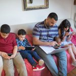 Pai e duas crianças usam dispositivos digitais sentados em um sofá, em casa.