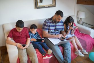 Pai e duas crianças usam dispositivos digitais sentados em um sofá, em casa.