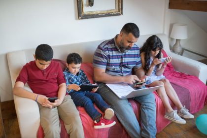 Pai e duas crianças usam dispositivos digitais sentados em um sofá, em casa.