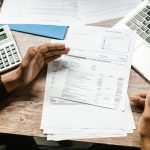 Mãos analisam documentos financeiros ao lado de uma calculadora e um laptop sobre uma mesa de escritório.