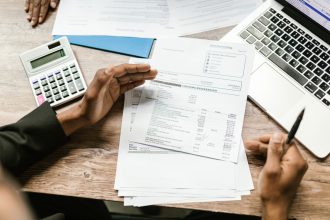 Mãos analisam documentos financeiros ao lado de uma calculadora e um laptop sobre uma mesa de escritório.